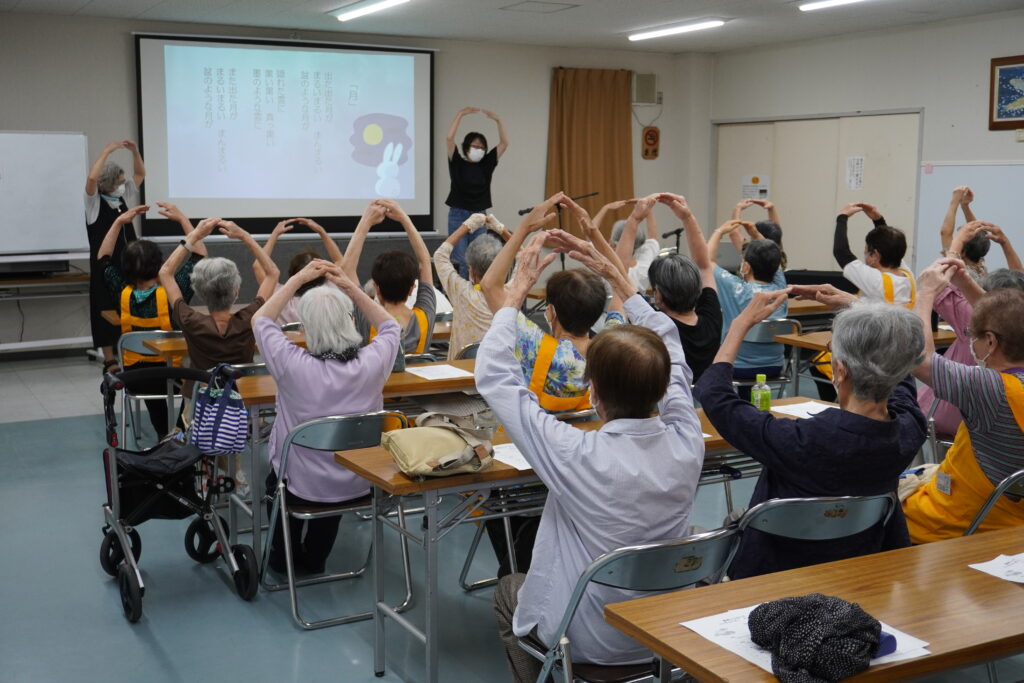 「ふしぎなたまご」さんがスクリーンの横でポーズを取り、会員さんがそれを真似して体操をしている