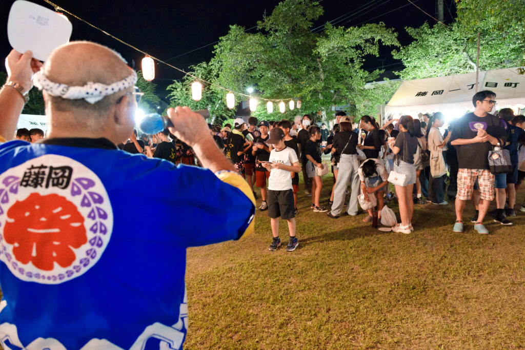 抽選番号入りのうちわを手に説明するふれあい部長と、それを聞く参加者たち