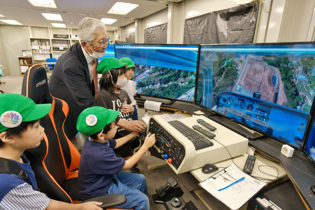 航空資料館の方の開設を受けながら、フライトシミュレーターを操作する子どもと、周りで見守る子どもたち