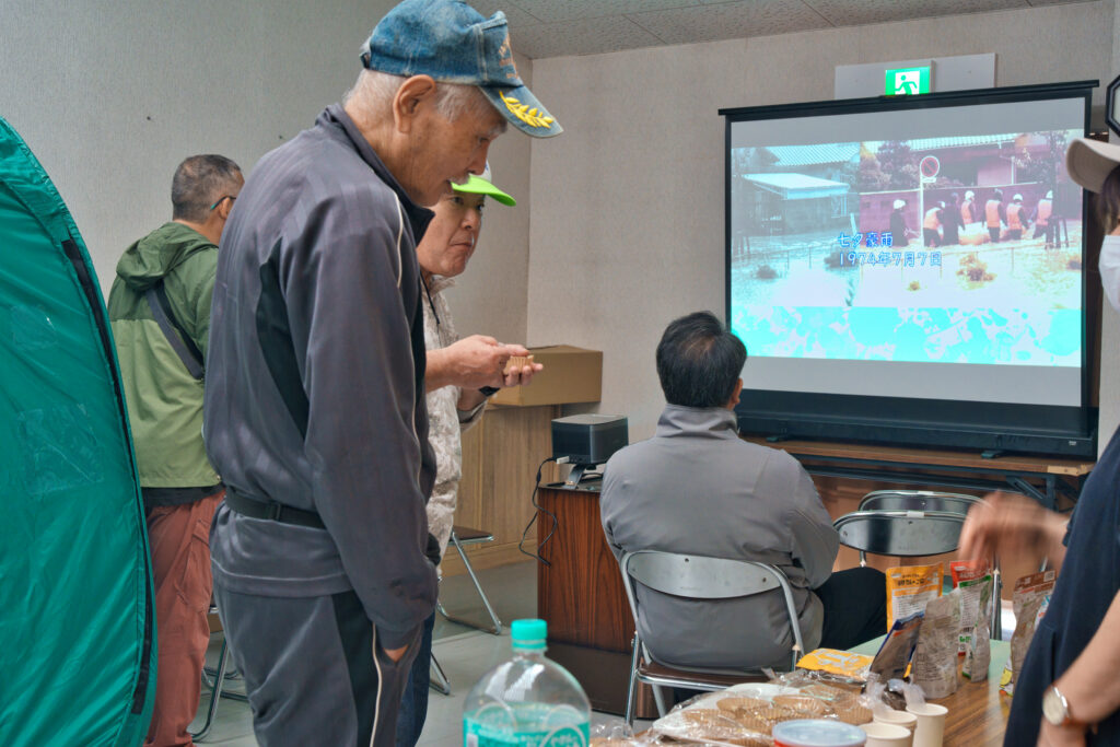防災食の試食をする住民の皆さんと、その背後で上映されている藤岡で発生した災害の映像を見る住民の皆さん