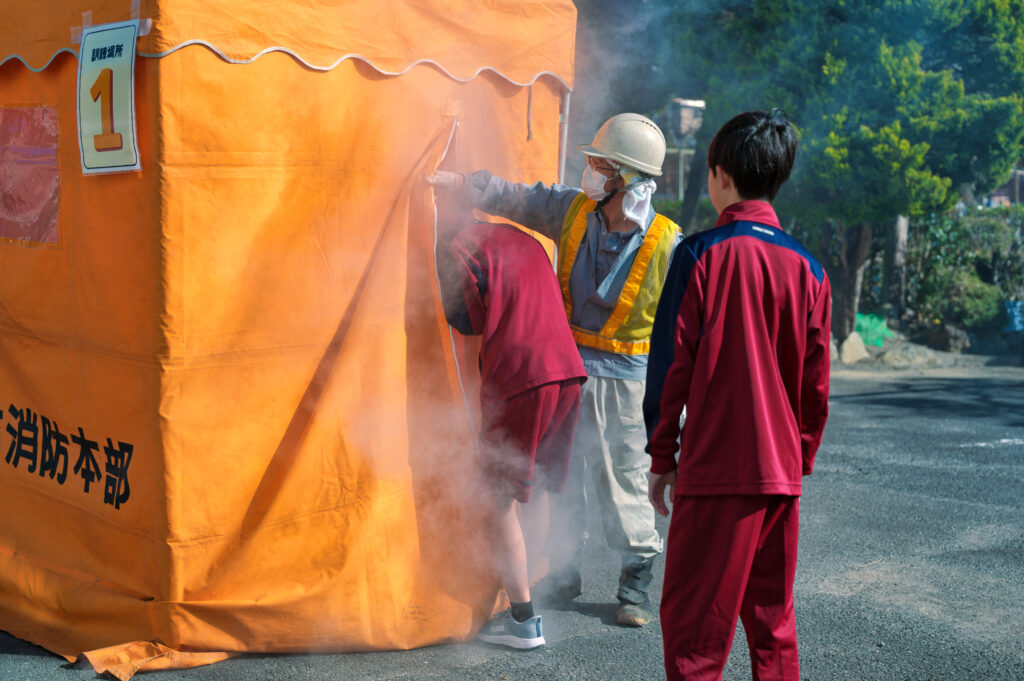 煙が充満したスモークハウスに入っていく中学生と、それを見守る中学生と役員