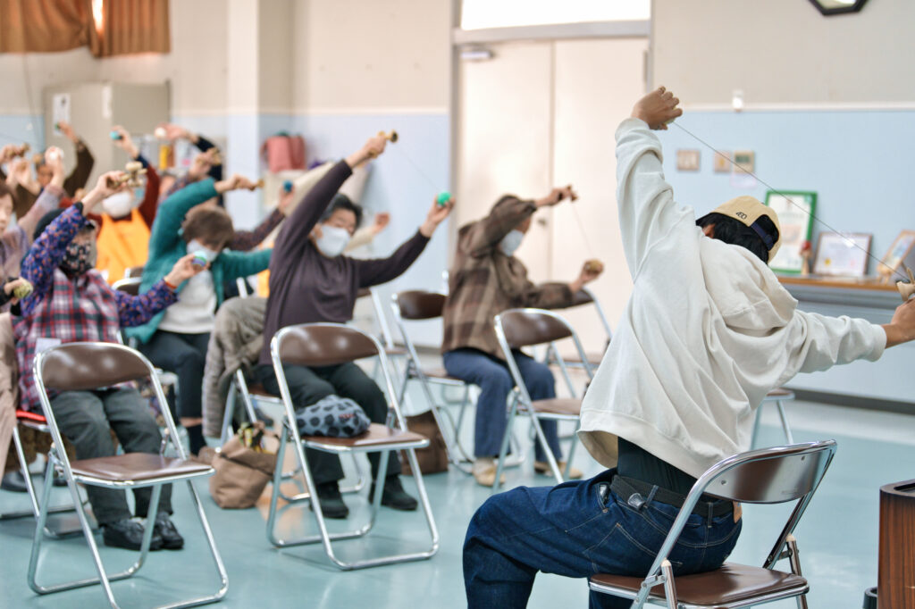 けん玉と球を別々の手で持ち、頭の上にあげ、手を広げ、体を倒す運動をしているぎんちゃんと会員たち