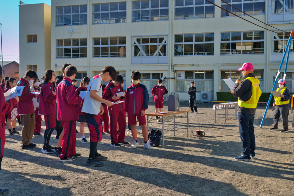グラウンドで役員から訓練の説明を受ける小中学生