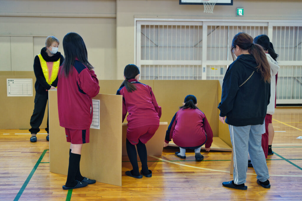 段ボール製簡易間仕切りを組み立てる小中学生たちと見守る役員たち