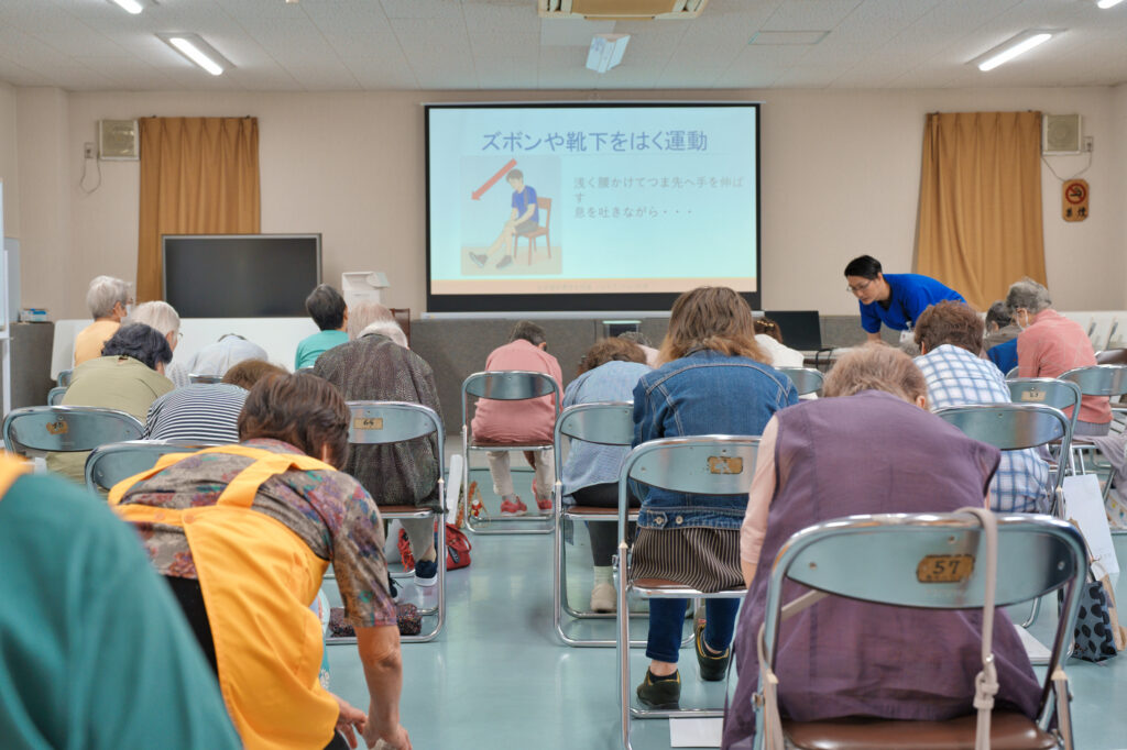 スクリーンに説明が映し出され、靴下を掴み上に上げる運動をする会員さんたちの後ろ姿