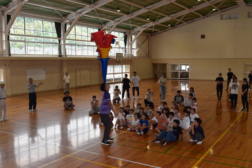 キリン先生が3個のポールの尖った方を頭に乗せて、それを見守る子どもたち