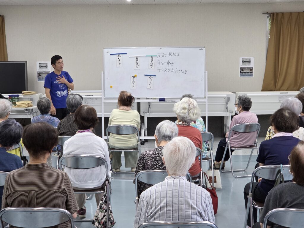 塚本先生がホワイトボードの前に立ち、会員たちに説明している